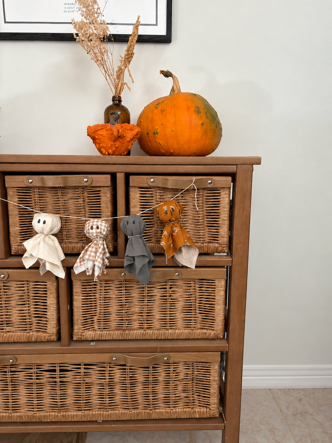 Garland of four fabric ghosts hanging across a wooden cabinet with wicker baskets, topped with two pumpkins and a vase of dried flowers.