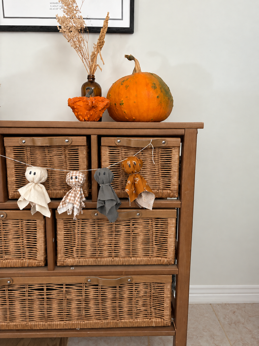 Garland of four fabric ghosts hanging across a wooden cabinet with wicker baskets, topped with two pumpkins and a vase of dried flowers.