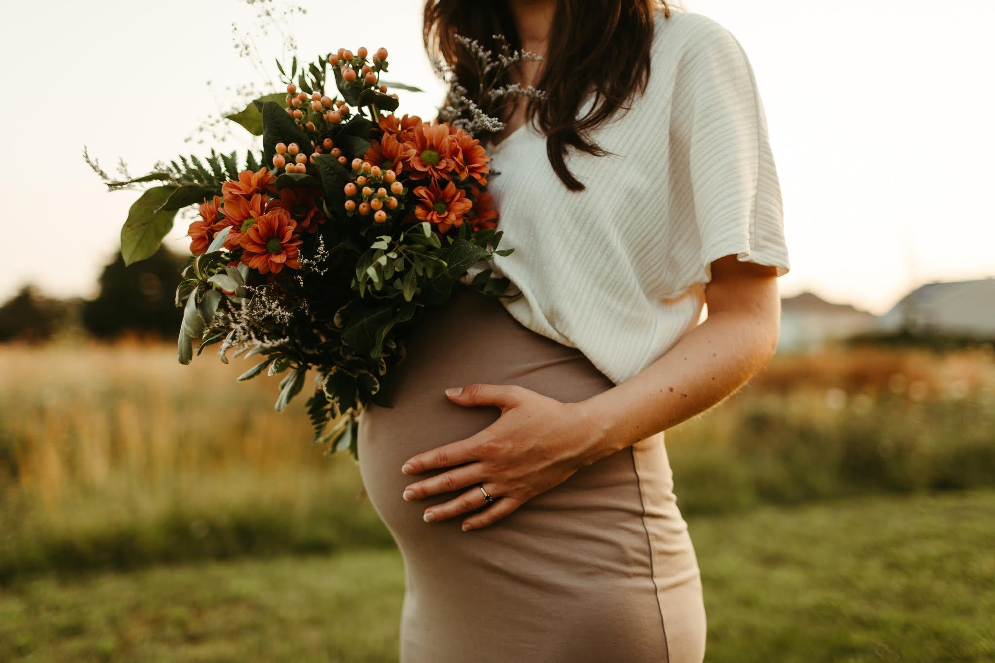 A pregnant woman in a white short-sleeve top and beige skirt holds a large autumn bouquet of orange flowers and green leaves, standing in a grassy field at sunset.