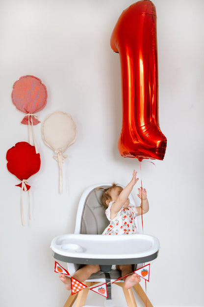 A baby sitting in a high chair reaches for a large metallic red number 1 balloon against a white wall. Three fabric balloon decorations in red, and cream hang on the left, and small red-and-white checkered bunting is attached to the high chair.
