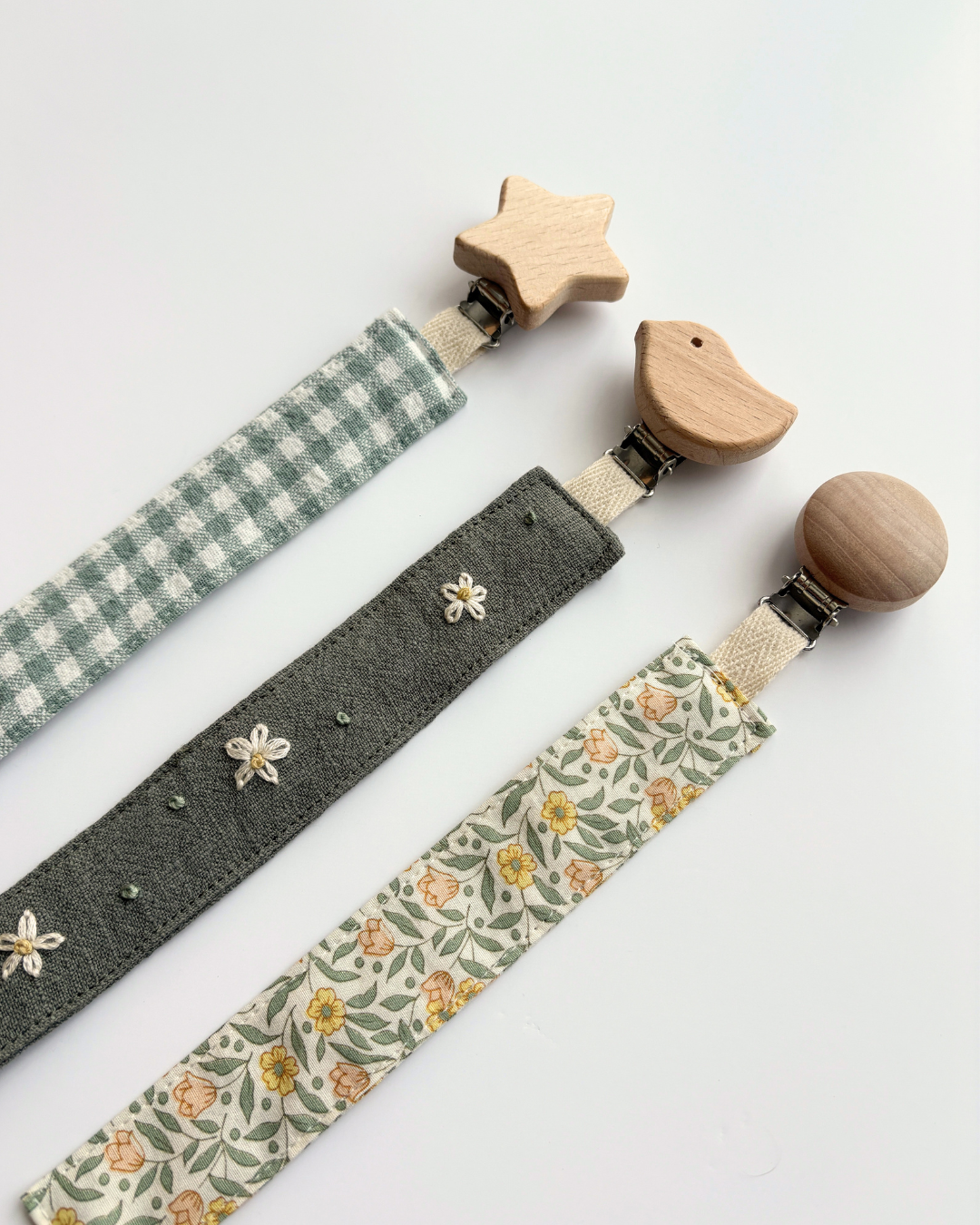 Three fabric baby pacifier clips with wooden animal and star-shaped clamps, laid out diagonally on a white background. The clips feature three different fabric designs: a green and white gingham check, a dark gray fabric with white embroidered flowers, and a cream floral print with yellow and orange flowers and green leaves.