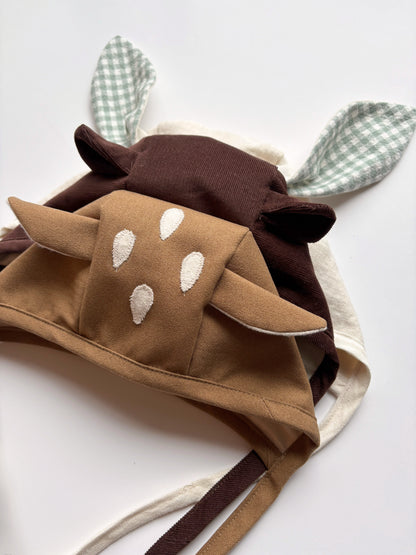 A soft fabric animal-themed bonnets in tan, brown and, with white embroidered spots, attached long ear-like tabs, and green-and-white gingham pointed ears, photographed on a plain white background.