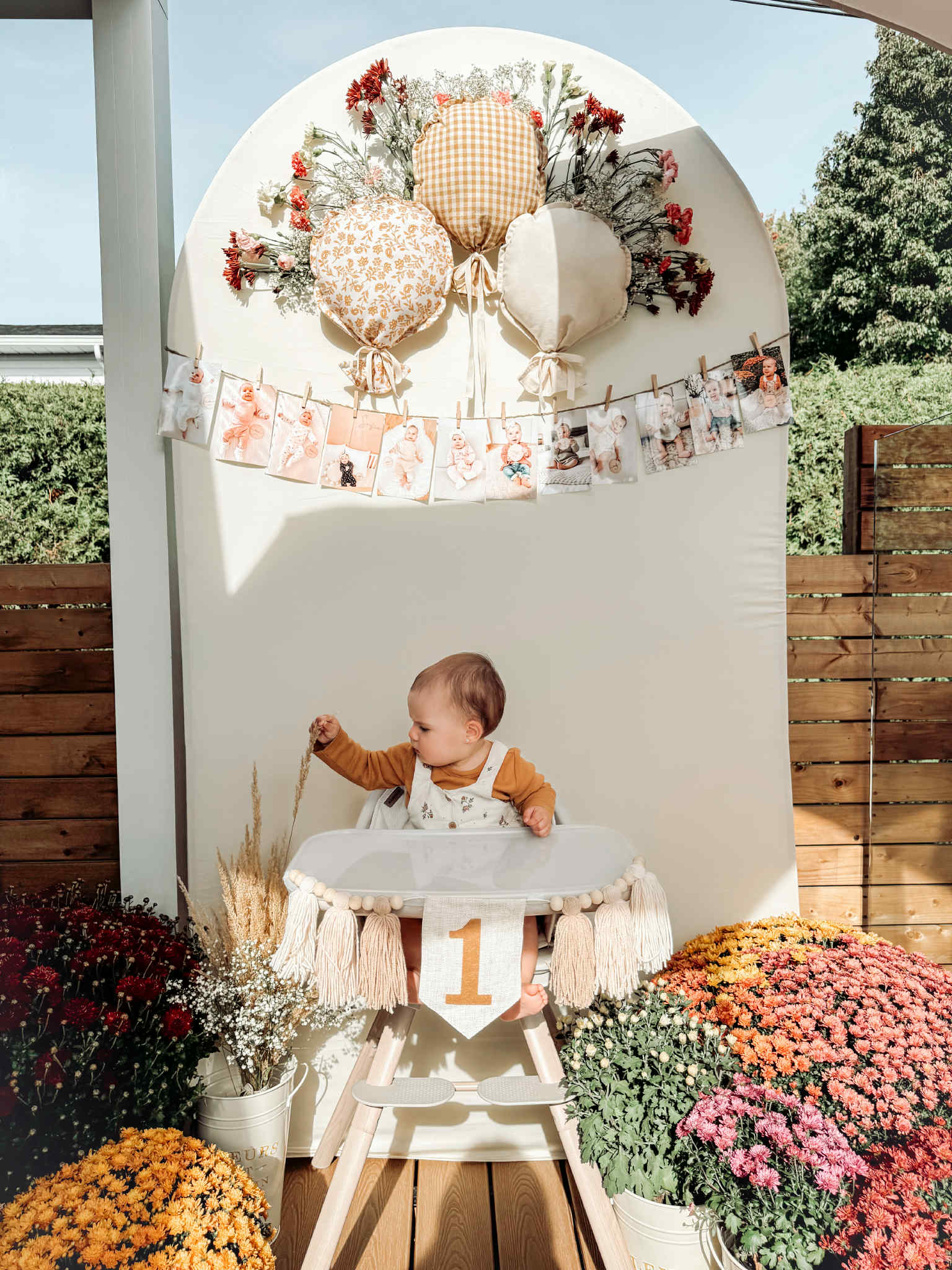 A baby sits in a decorated high chair under a white arched backdrop with balloon decorations, a photo banner, and colorful flower arrangements. The baby is wearing a brown top and white overalls.