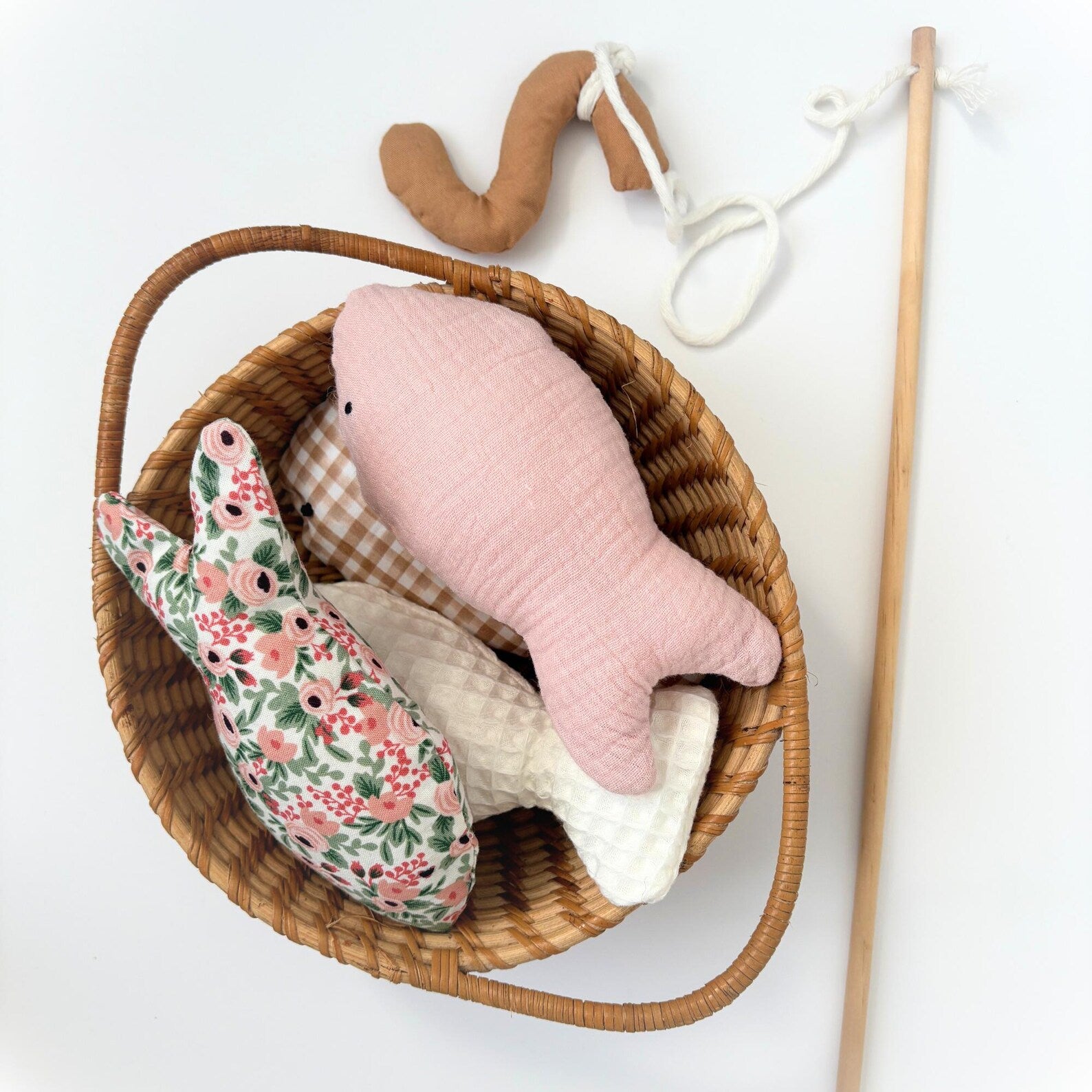 A styled flat-lay of a woven wicker basket containing a pink quilted fish, a floralfish, and a white gingham fish, with a brown worm toy and a wooden stick attached to it on a white background.