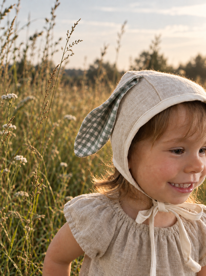 A young child wearing a beige linen bonnet with green-and-white gingham bunny ears, standing in a sunlit grassy field.