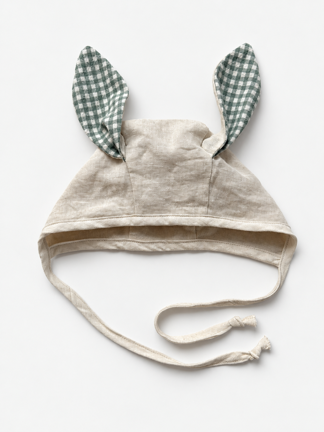 A beige linen baby bonnet with long ties and two upright bunny ears made of green and white gingham fabric, shown on a plain white background.