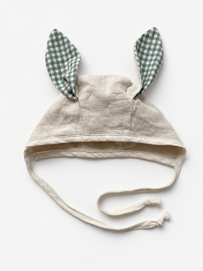 A beige linen baby bonnet with long ties and two upright bunny ears made of green and white gingham fabric, shown on a plain white background.