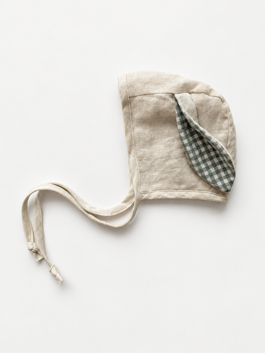 A light beige linen bonnet with a green and white gingham patterned ear flap, laid flat on a white background.