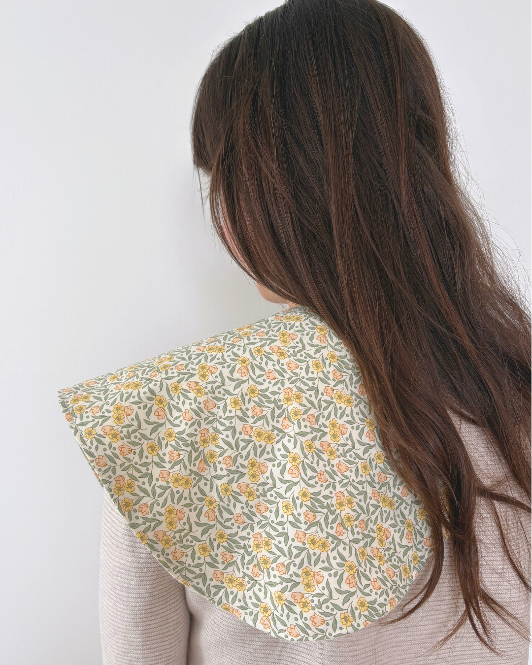 A close-up back view of a person with long dark brown hair wearing a wide, flared burpcloth with a delicate yellow and orange floral pattern on a light sage green background.