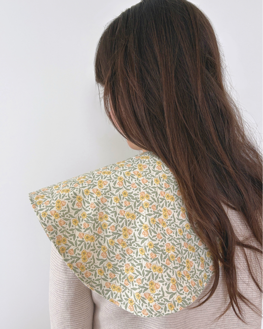 A close-up back view of a person with long dark brown hair wearing a wide, flared burpcloth with a delicate yellow and orange floral pattern on a light sage green background.