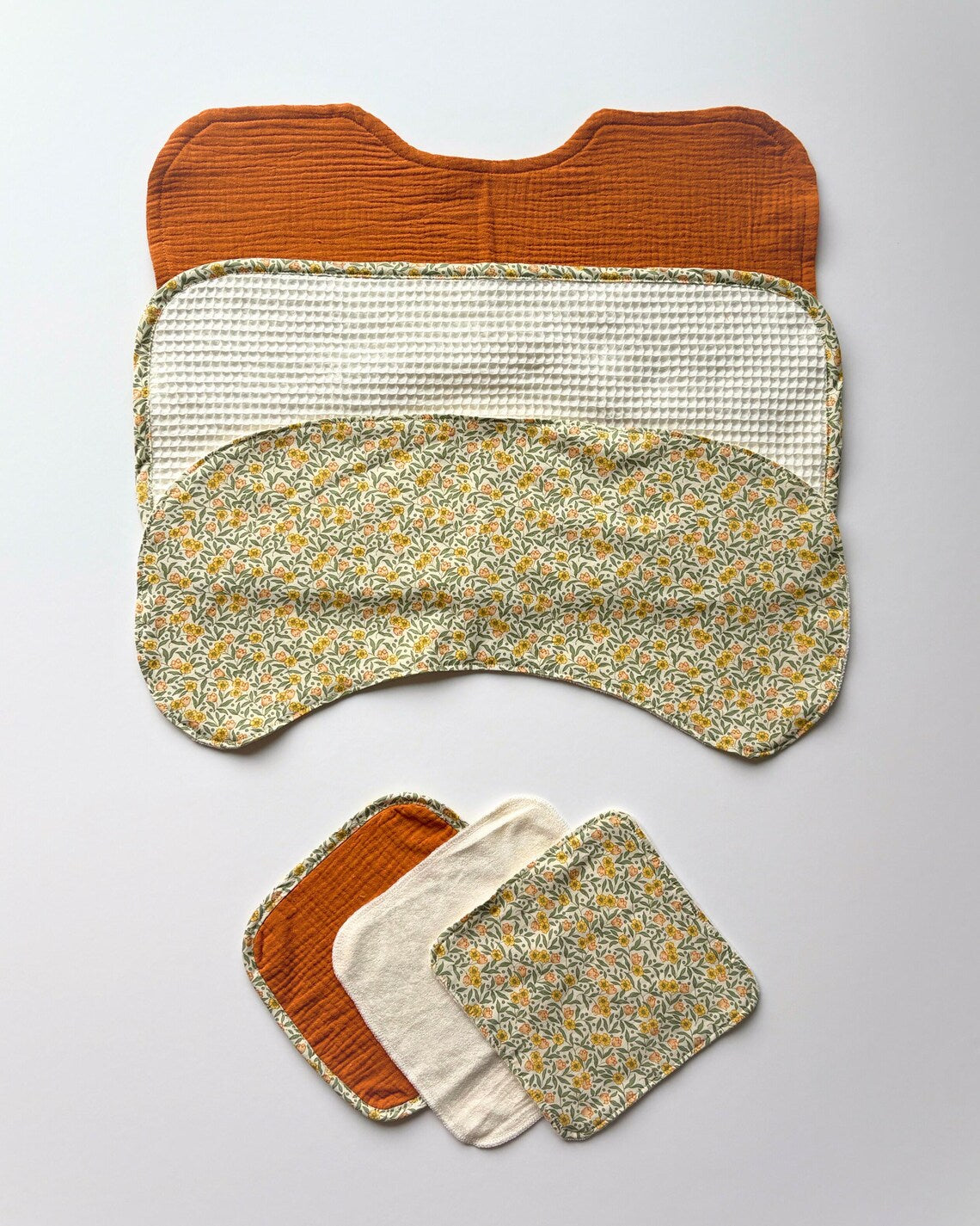 Three reusable burpcloths are arranged on a light gray background: one rust orange ribbed cloth, one cream waffle cloth, and one cream-and-yellow floral patterned cloth. A smaller set of three matching washcloths is stacked below.