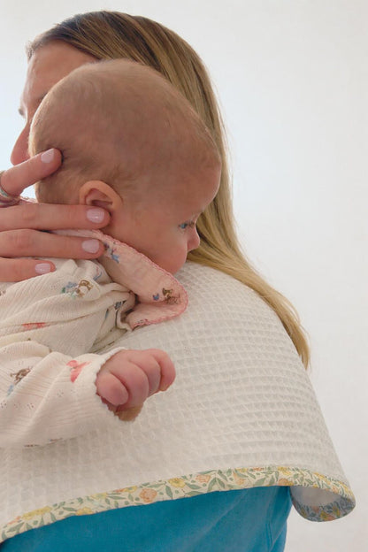 A close-up lifestyle shot of a woman holding a baby against her shoulder. The baby is wearing a cream-colored textured waffle-weave onesie with a pink collar, and the woman is wearing a blue top with a matching cream waffle-weave baby burp cloth draped over her shoulder.
