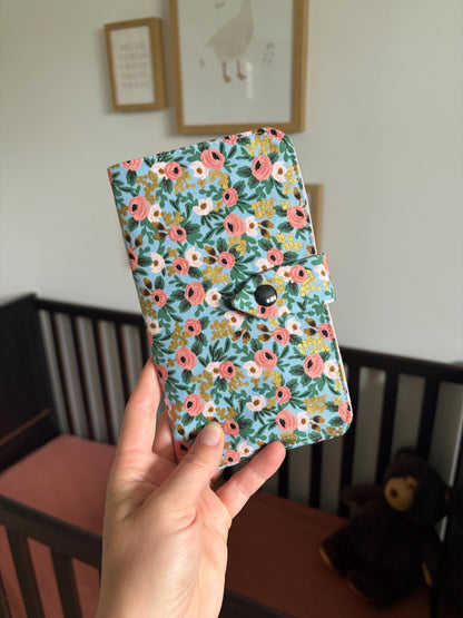 A hand holding a floral-patterned health record cover with a snap button closure, set against a softly blurred nursery background with a crib and framed wall art.