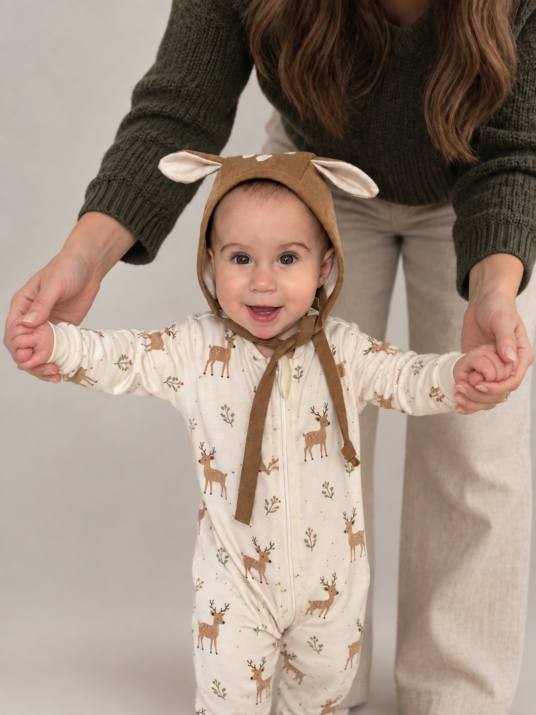A baby wearing a cream-colored footed sleeper with a brown deer print and a matching brown bunny-ear bonnet, being supported by an adult in a dark green sweater and beige pants against a neutral studio background.