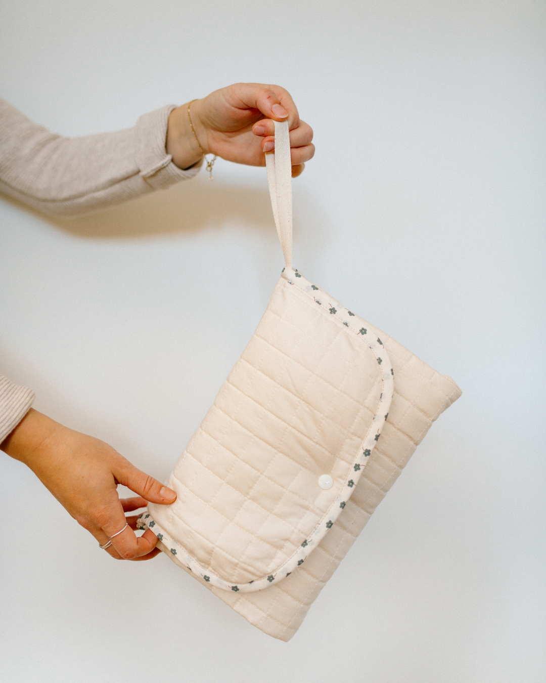 A person holds a cream-colored quilted travel changing mat with a matching fabric strap. The bag features a quilted texture, a snap-button closure, and a decorative border of small blue flowers.