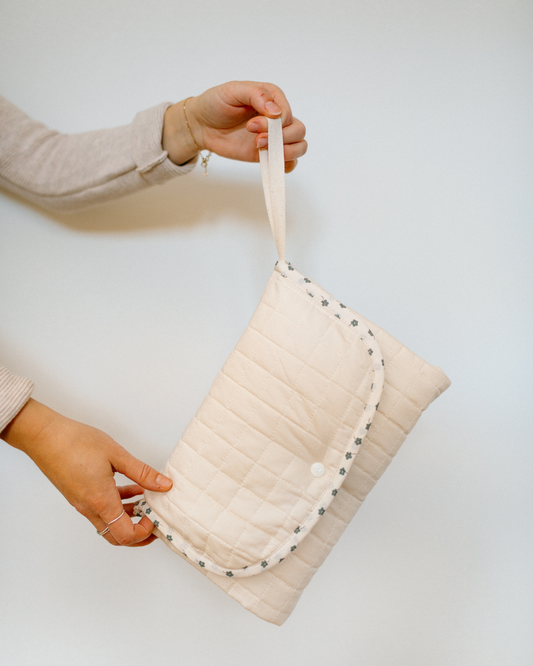 A person holds a cream-colored quilted travel changing mat with a matching fabric strap. The bag features a quilted texture, a snap-button closure, and a decorative border of small blue flowers.