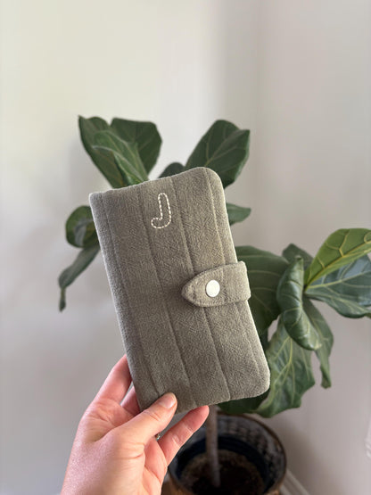 A hand holding a sage green fabric wallet or planner cover with a snap button closure and white embroidered initials, set against a background of a green potted plant and a light-colored wall.