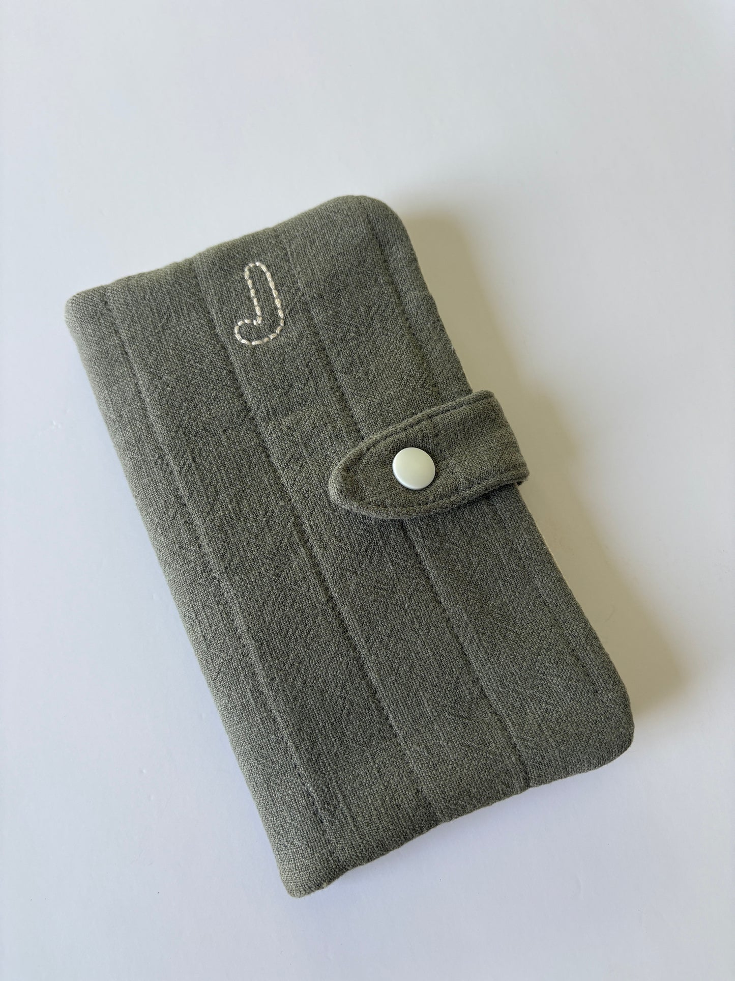 An olive green textured fabric pouch with a snap-button closure and a small white embroidered letter 'J' on the front, shown on a plain light background.