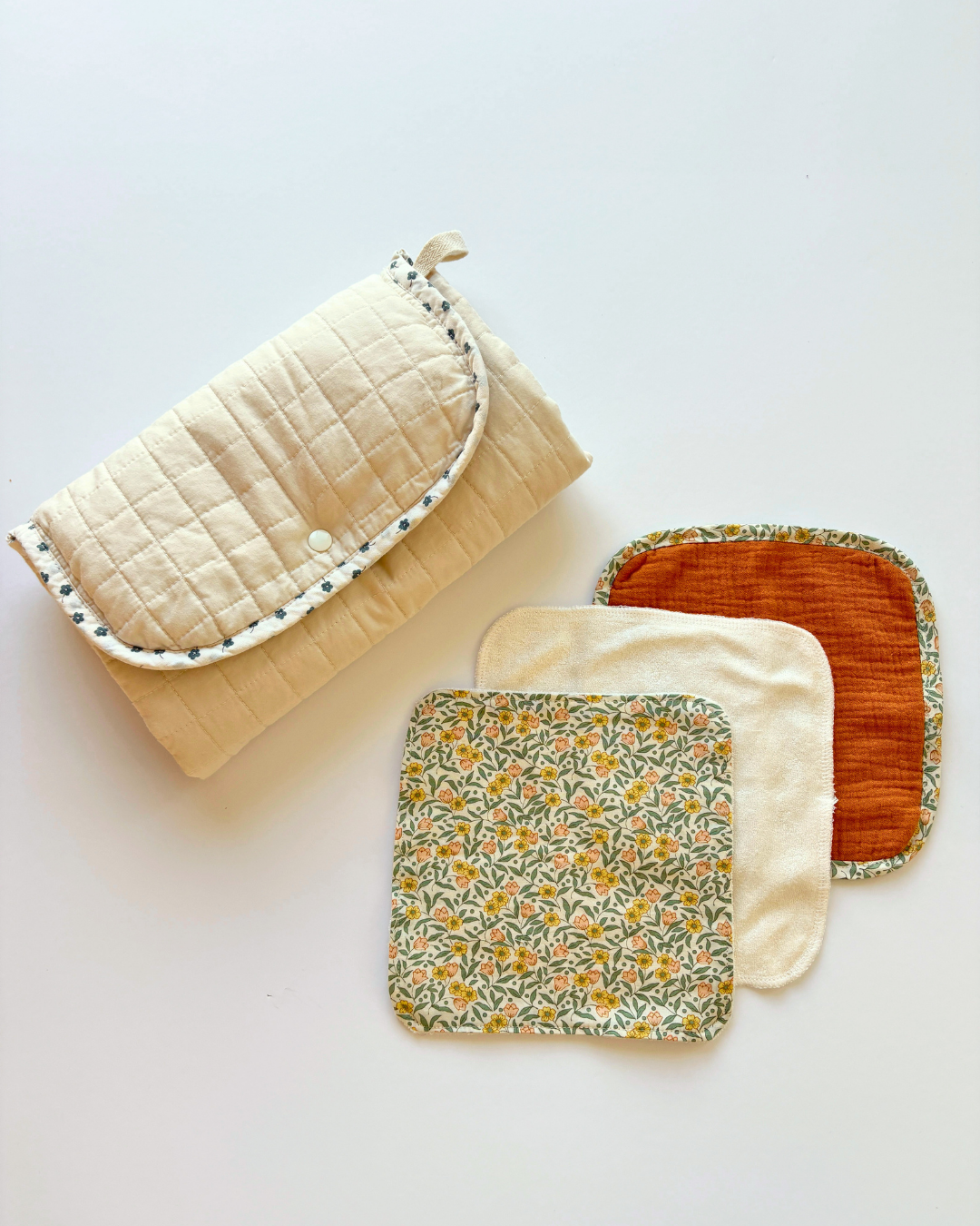 A quilted cream fabric travelc changing mat with a floral trim, displayed next to three square fabric washcloths in cream, orange corduroy, and a yellow floral pattern, arranged on a plain white background.