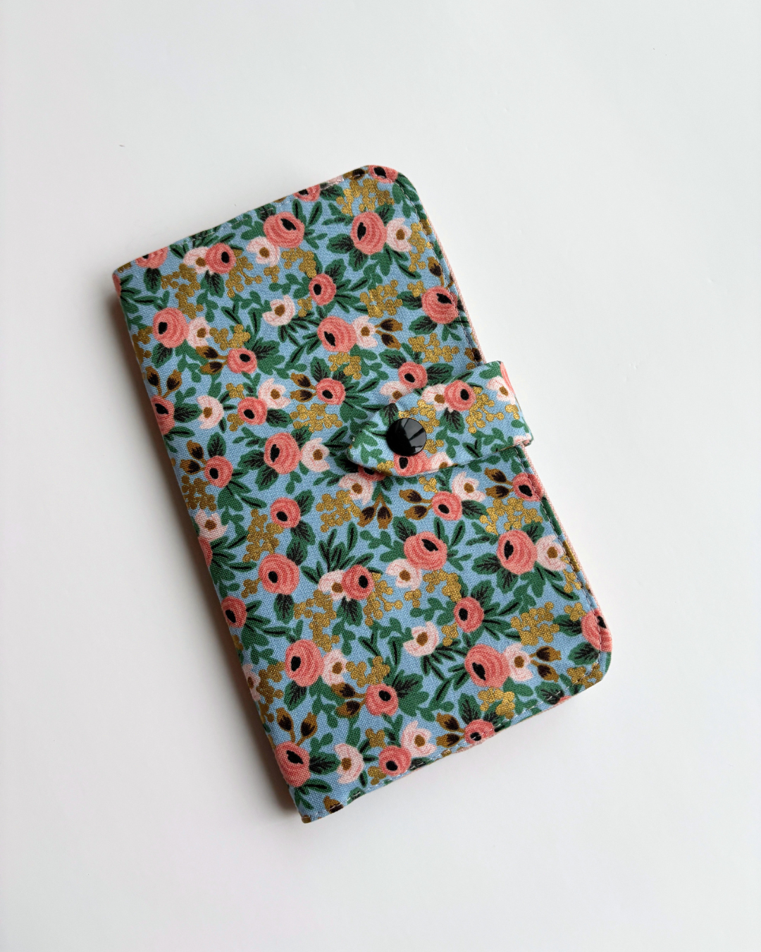 A fabric health record cover with a dense floral pattern in pink, green, yellow, and white on a light blue background, featuring a black snap-button closure.