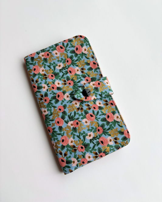 A fabric health record cover with a dense floral pattern in pink, green, yellow, and white on a light blue background, featuring a black snap-button closure.