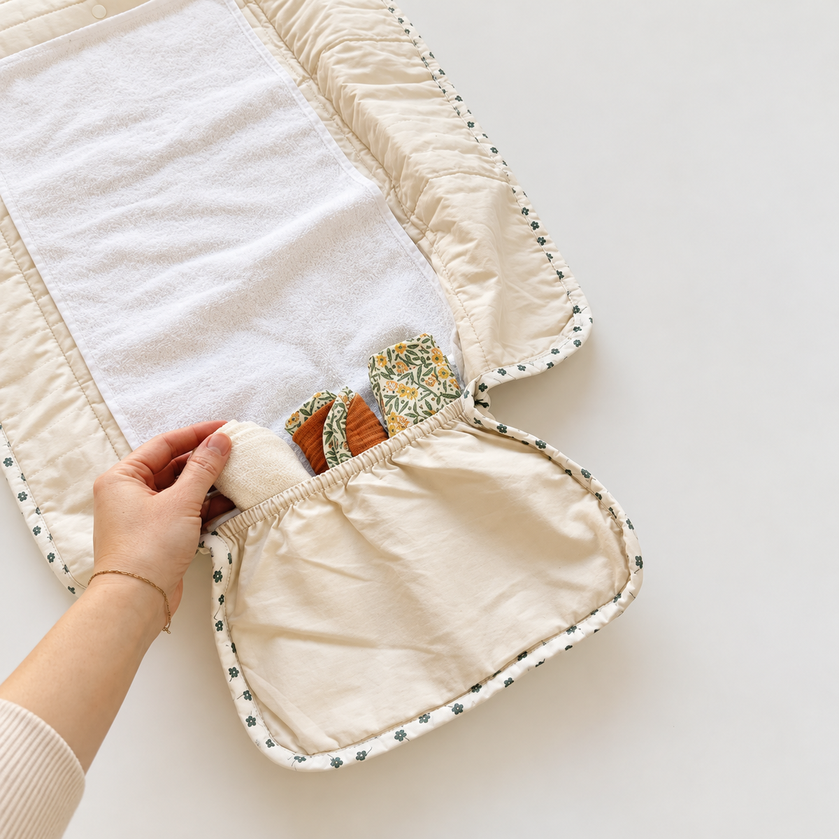 A close-up of a beige quilted baby travel changing pad with a white insert, shown on a light neutral background. A hand is pulling open a front pocket that contains small patterned fabric accessories.