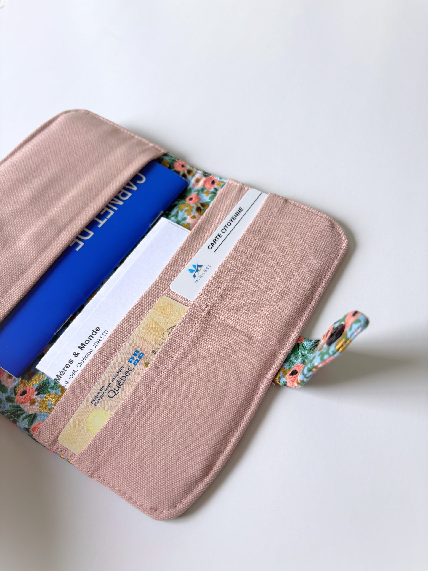 A blush pink canvas organizer pouch shown open on a white background, containing various cards and a blue vaccination book, with a floral-patterned interior lining and strap.