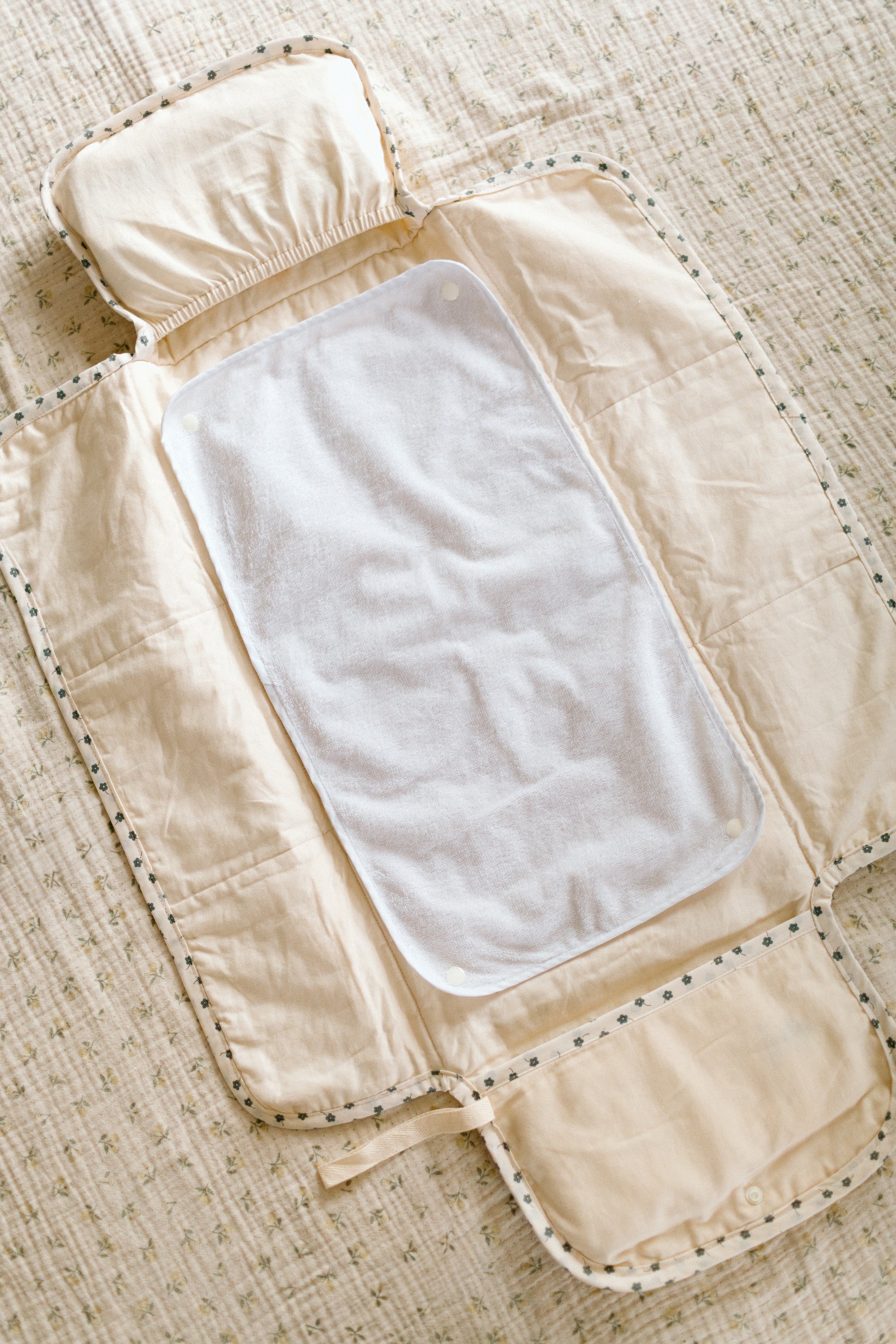 A cream-colored quilted baby changing mat with a white pad liner, laid flat on a textured beige flowered double gauze blanket.