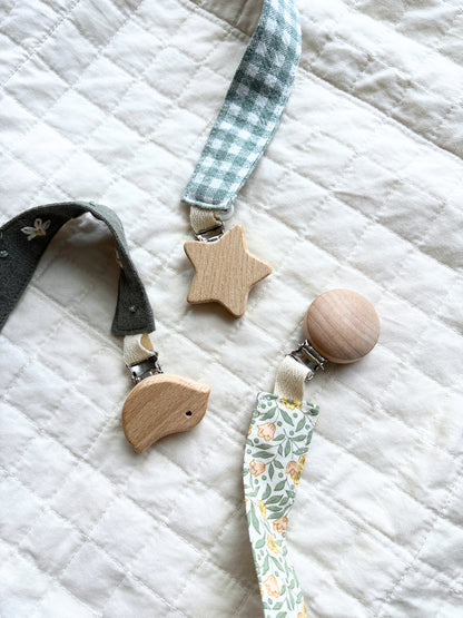 Three fabric pacifier clips with wooden accents are arranged on a white quilted blanket. One clip has a dark green strap and a light wood bird-shaped clip.