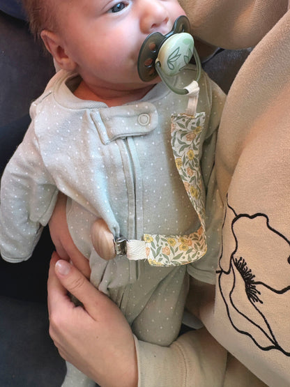 A close-up lifestyle shot of a baby wearing a light sage green dot one-piece romper with a floral-patterned pacifier clip and a wooden spoon clasp, held by an adult wearing a beige sweatshirt.