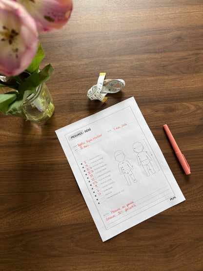 A printed sizing/measurement sheet for a baby or child's sleep hooder lies on a wooden tabletop, styled with a pink flower vase, a tape measure, and a coral pen nearby.