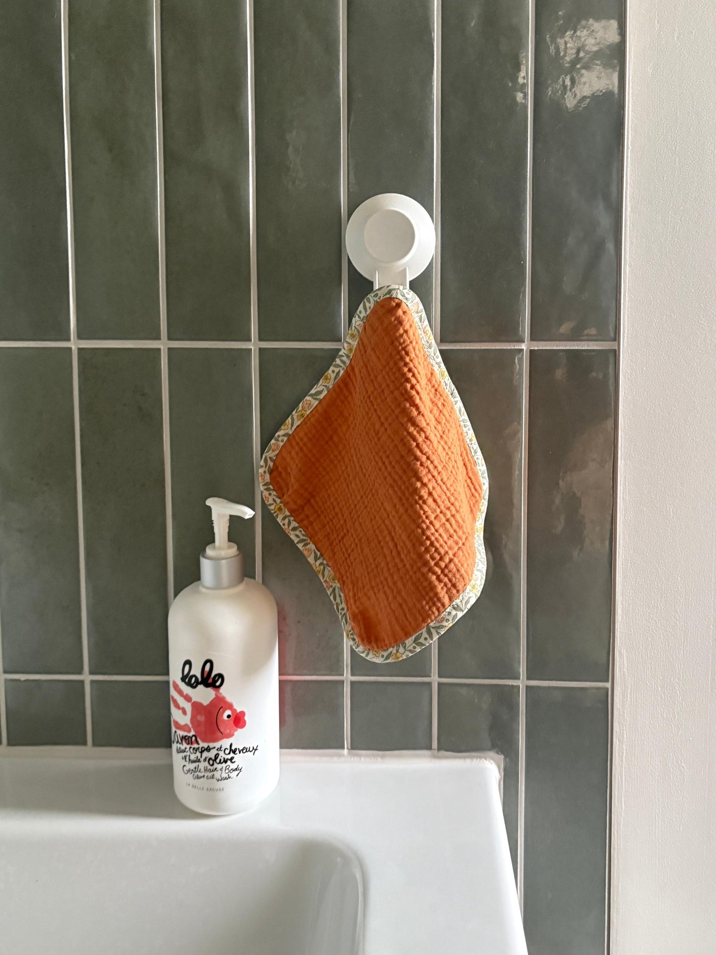 An orange textured washcloth with a patterned border hangs from a white suction cup on a dark green tiled bathroom wall above a white sink, next to a bottle of hand wash.