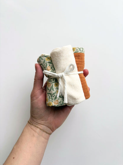 A hand holding a small bundle of three folded cloth items tied with a white string, including a green floral patterned piece, an off-white textured piece, and a rust-orange piece, against a plain light background.
