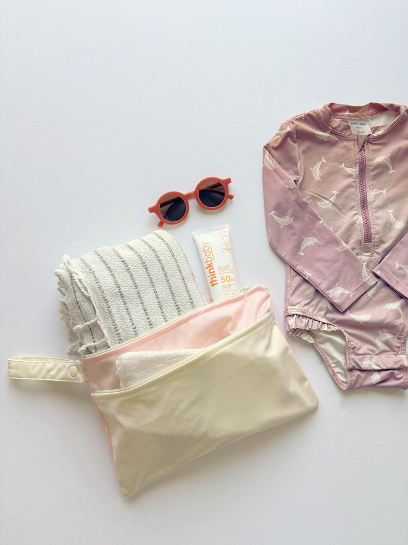 A styled flat-lay of wet bag on a white background, including a dusty pink long-sleeve baby bodysuit with white dolphin prints, a cream and pink zippered pouch, a striped swaddle, a tube of thinkbaby Sunscreen, and orange-framed sunglasses.