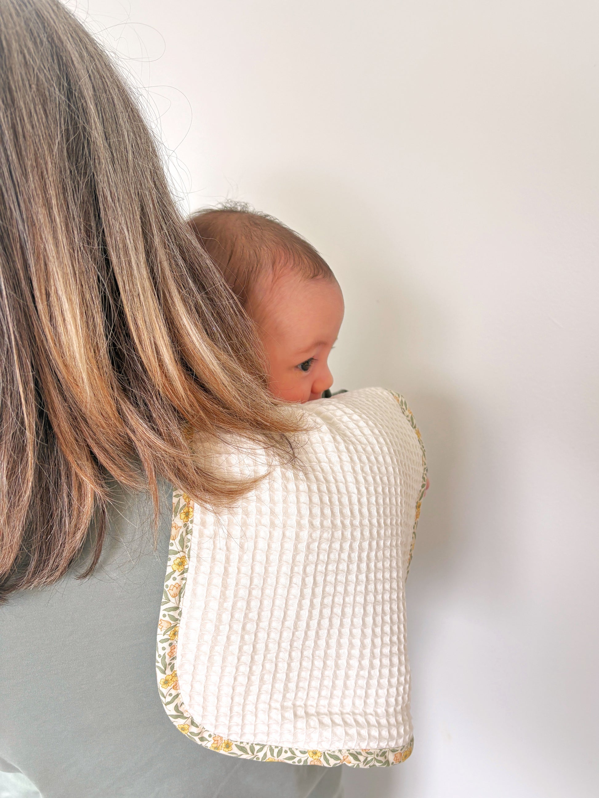 A baby peeking out from behind a woman's shoulder wearing a waffle-knit burp cloth with a yellow and green floral bias binding.