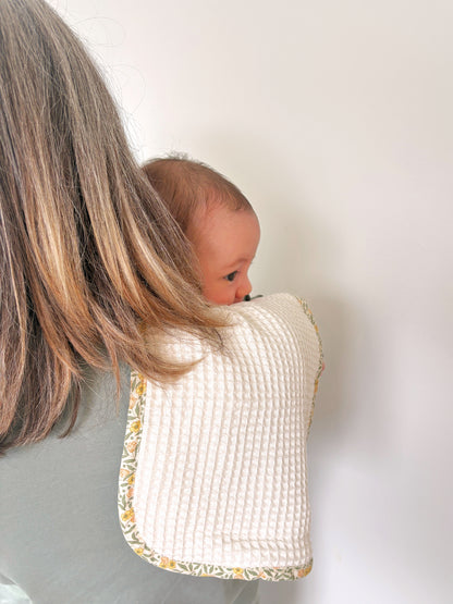 A baby peeking out from behind a woman's shoulder wearing a waffle-knit burp cloth with a yellow and green floral bias binding.