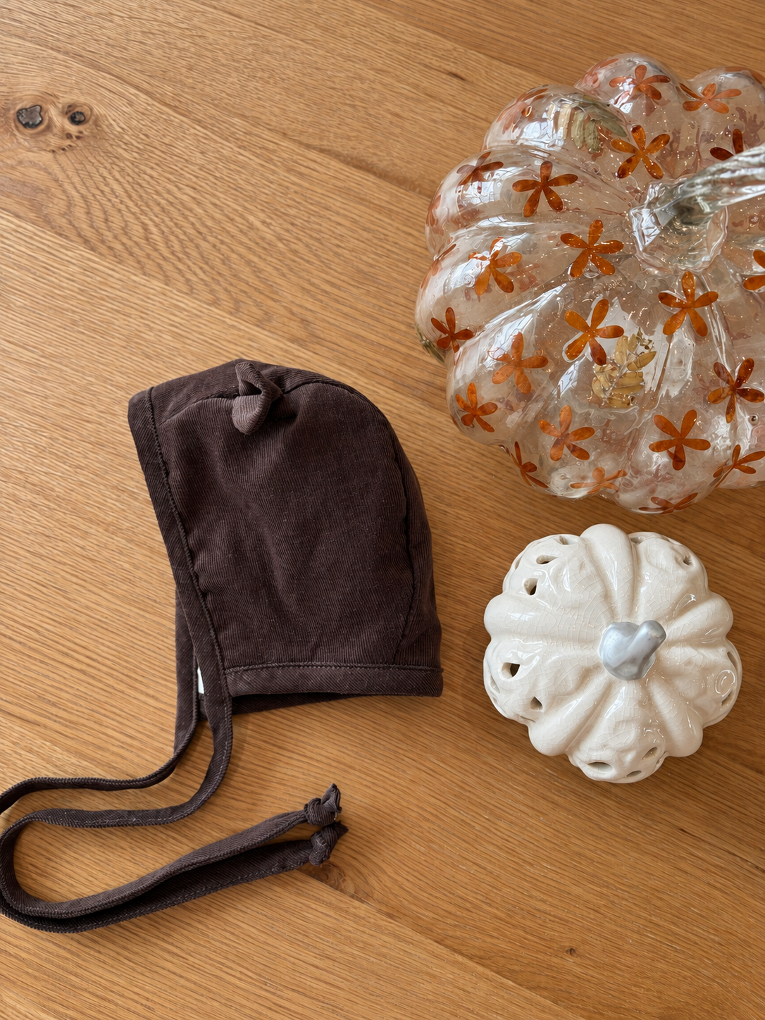A dark brown fabric bonnet with adjustable ties is displayed on a light wood surface next to decorative glass pumpkins and a white ceramic pumpkin.