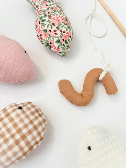 A set of handmade fabric animal toys arranged on a white background, including a pink whale, a floral fish, a tan worm, a brown gingham fish, and a white fish, with a wooden stick and white string visible.