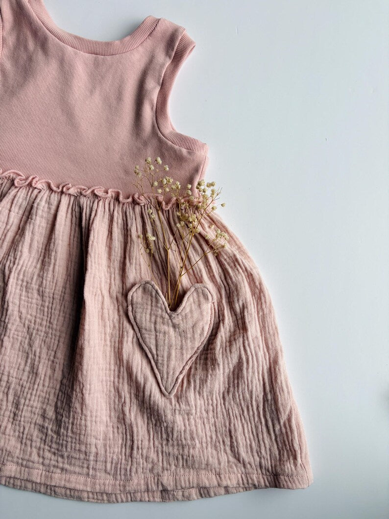 A dusty pink sleeveless baby dress made of textured crinkled gauze, featuring a solid ribbed top and a gathered skirt with a large applique heart pocket holding dried baby's breath flowers, shown against a light gray background.