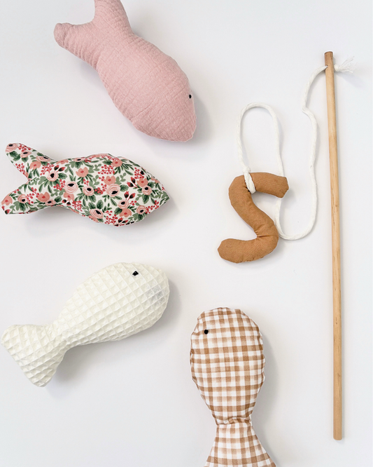 A set of handmade fish-shaped and a worm fabric toys and a wooden fishing rod arranged on a clean white background. The toys feature various fabric patterns including solid pink, floral green and pink, textured cream, and brown gingham, with one toy attached to a white loop and the fishing rod.