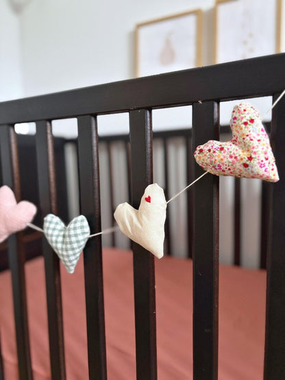 A string of handmade fabric heart garlands hanging across a dark wooden crib. The hearts are made from different fabrics: solid pink, green-and-white gingham, cream with a tiny red heart, and a pink, yellow, and white floral print.