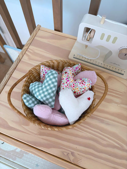 A collection of handmade fabric heart-shaped pillows in various patterns (checkered, floral, solid pink, white with a red heart) arranged in a small woven basket on a wooden table next to a miniature sewing machine.