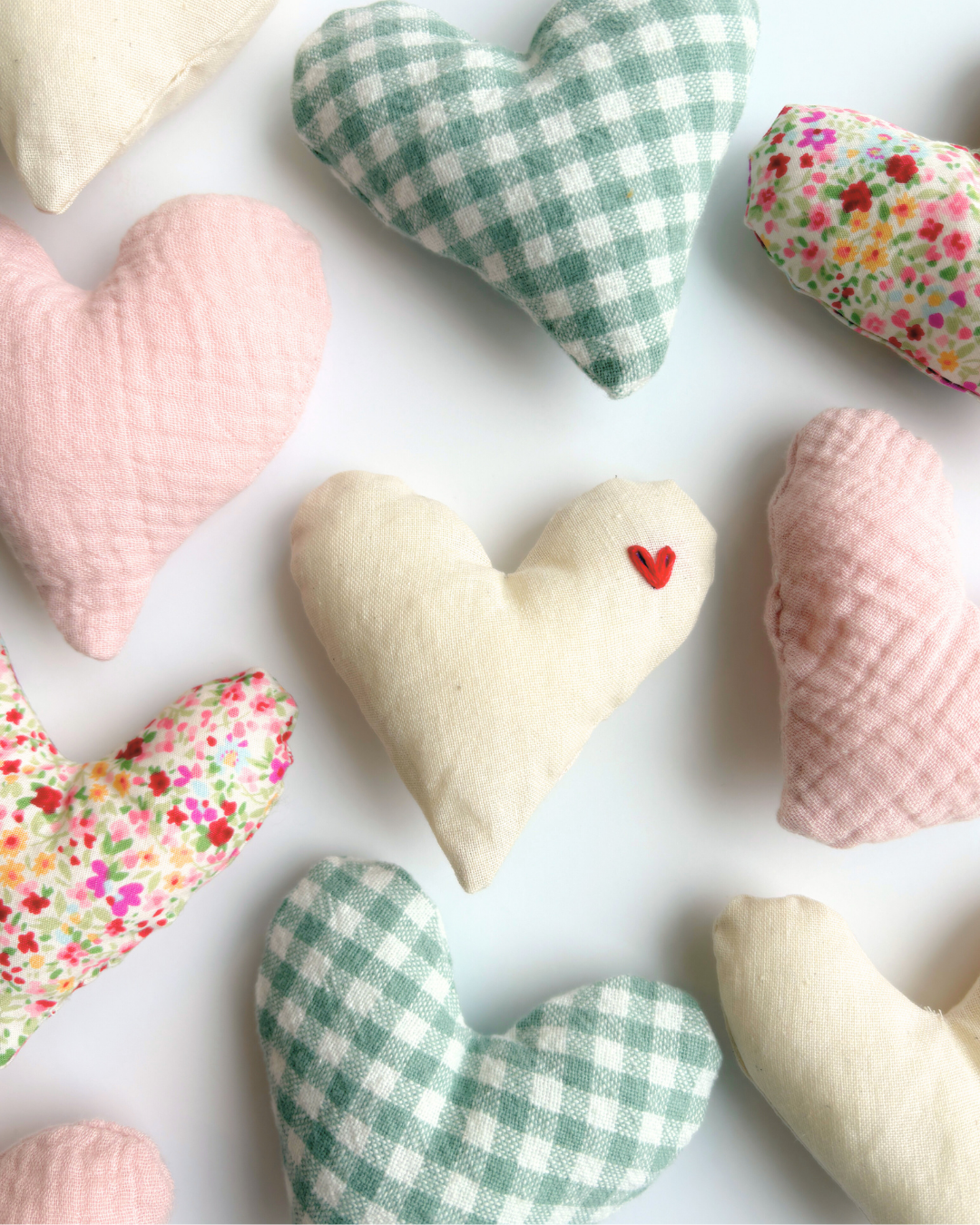 A collection of handmade fabric heart in various patterns and colors, including pink, green gingham, cream, and floral prints, arranged on a white background.