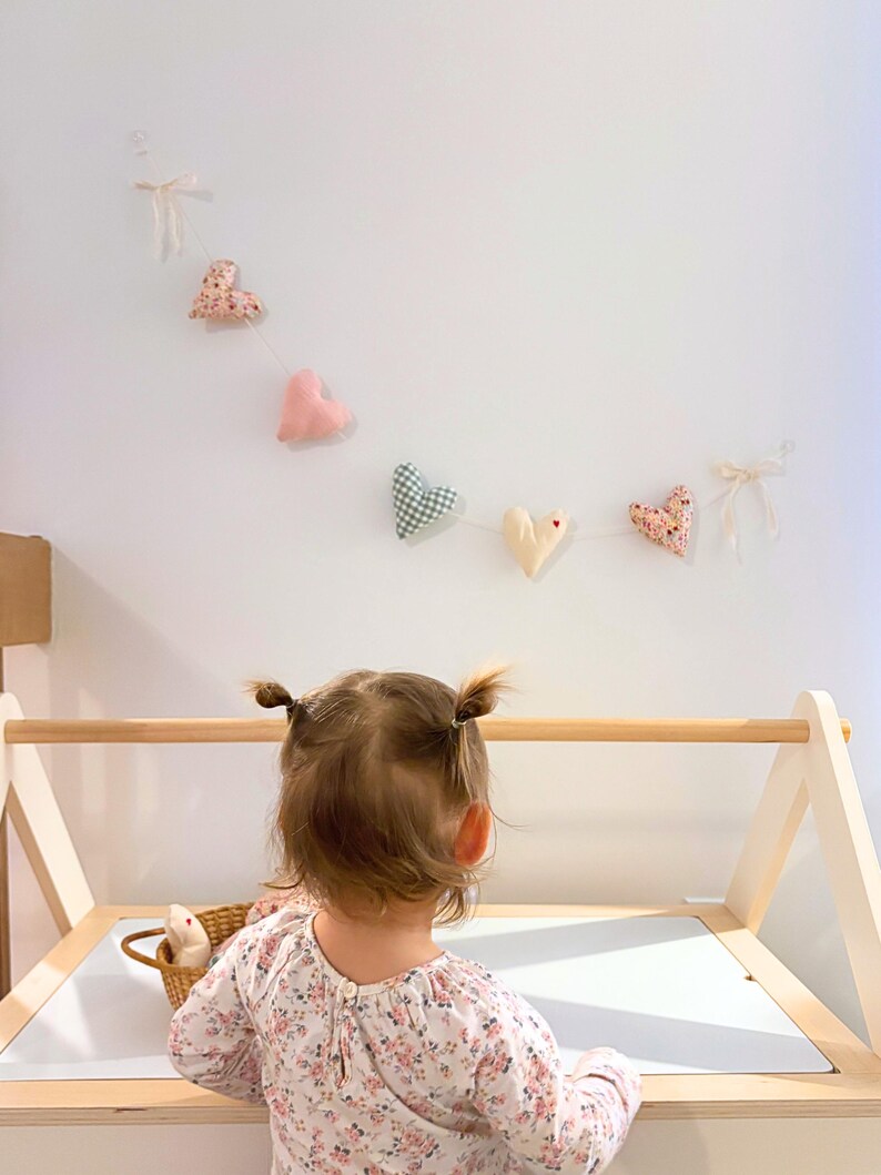 A toddler girl with two pigtails stands at a wooden activity table, looking up at a string of decorative fabric hearts hanging on the wall.