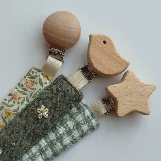 Video showing a close up of three fabric baby pacifier clips with wooden animal and star-shaped clamps, laid out diagonally on a white background. The clips feature three different fabric designs: a green and white gingham check, a dark gray fabric with white embroidered flowers, and a cream floral print with yellow and orange flowers and green leaves.