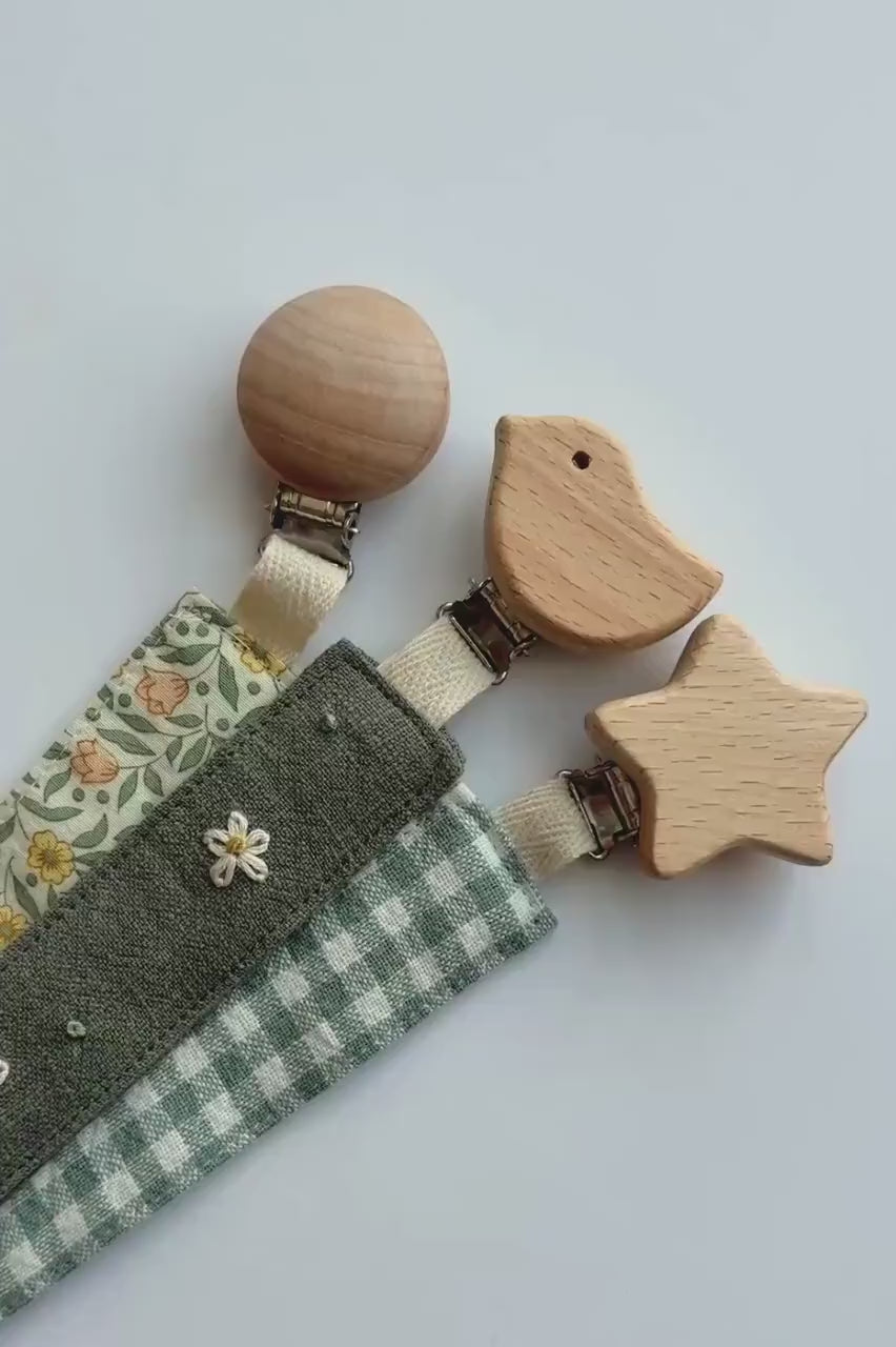 Video showing a close up of three fabric baby pacifier clips with wooden animal and star-shaped clamps, laid out diagonally on a white background. The clips feature three different fabric designs: a green and white gingham check, a dark gray fabric with white embroidered flowers, and a cream floral print with yellow and orange flowers and green leaves.