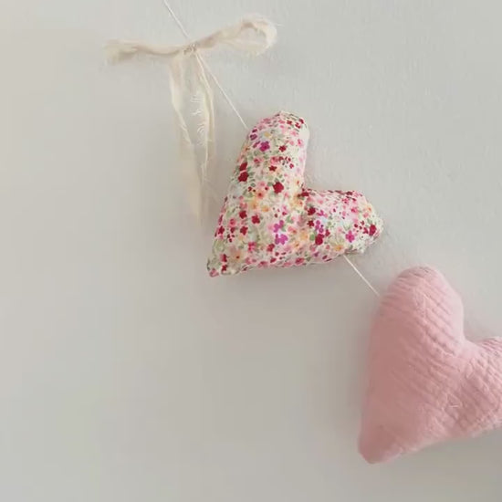 A video showing a garland of handmade fabric heart in various patterns and colors, including pink, green gingham, cream, and floral prints, arranged on a white background.