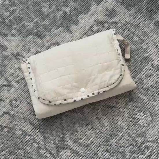 Video showing a beige quilted portable changing mat with a snap-button closure, two pockets, and a white removable liner inside.