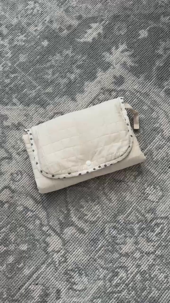 Video showing a beige quilted portable changing mat with a snap-button closure, two pockets, and a white removable liner inside.