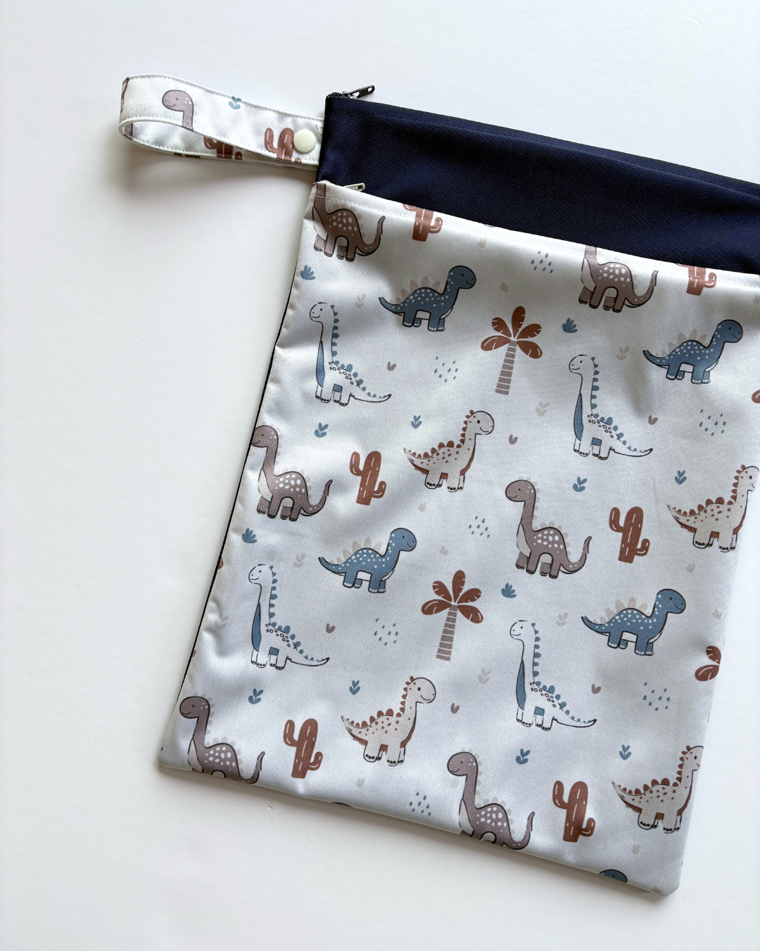 A fabric zippered wet bag with a dinosaur print, featuring a white background with brown, blue, and gray dinosaurs, a navy blue zipper track, and a matching wrist strap.